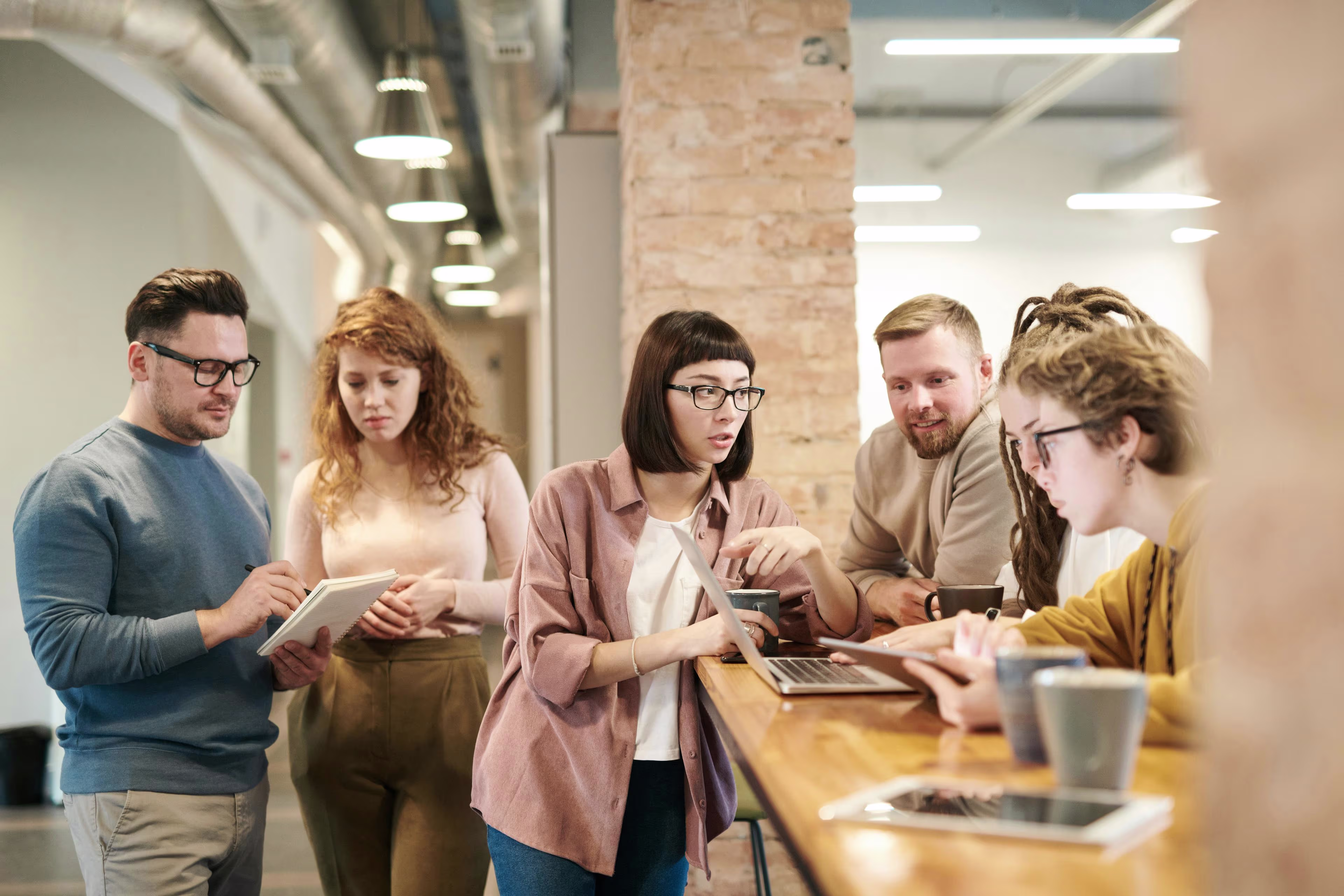 Group of people collaborating around a wooden table with laptops, notebooks, and coffee cups in a modern office.