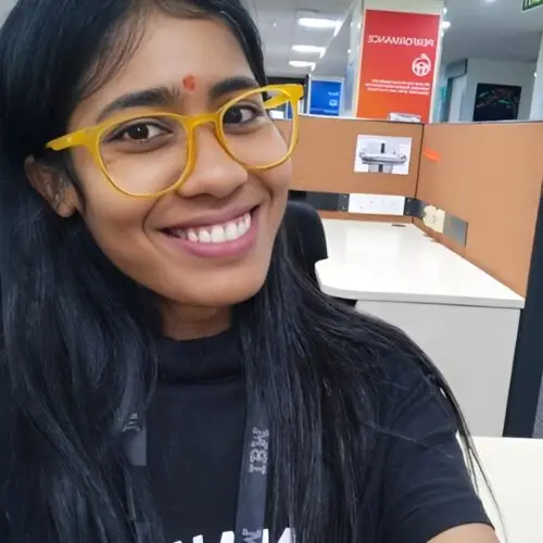 Smiling woman wearing yellow glasses and a black top taking a selfie in a modern office cubicle workspace.
